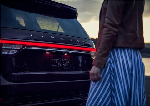 A person is shown near the rear of a 2023 Lincoln Aviator® SUV as the Lincoln Embrace illuminates the rear lights | Brinson Lincoln of Athens in Athens TX