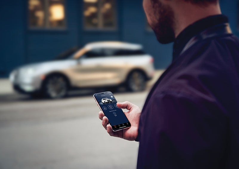 A person is shown interacting with a smartphone to connect to a Lincoln vehicle across the street. | Brinson Lincoln of Athens in Athens TX